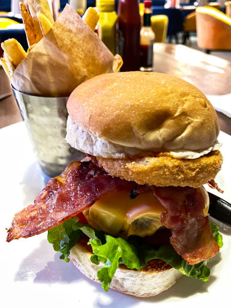 istock Close-up image of restaurant with gourmet double cheeseburger, bacon, melted cheese, tomatoes, lettuce and onion in seedless burger bun on white plate, portion of French fry chips served in a dimpled metal cup lined with greaseproof parchment paper, 1474120619