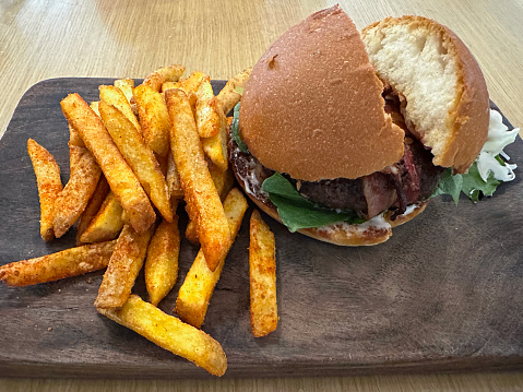 Stock photo showing close-up, elevated view of meal of bacon hamburger in seedless burger bun on wooden chopping board, served with crunchy French fry chips.
