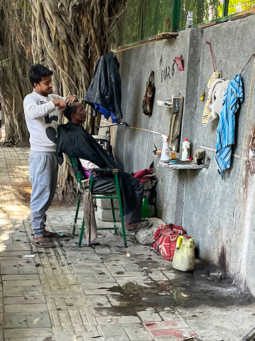 Delhi, India - December 9, 2022: Stock photo showing an outdoor, roadside barber's business with chair set up on pavement in front of mirror hung on concrete wall.