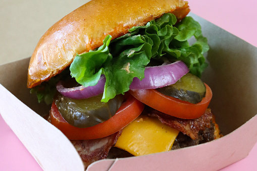 Stock photo showing close-up view of bacon cheeseburger served in a bread bun, in a cardboard takeaway box. The burger is made with a beef patty, bacon rasher, melted cheese, sliced tomato, red onion, pickled gherkin and lettuce.