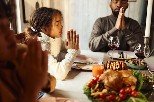 Religious African American girl praying before lunch with her single father and brother at dining table.
