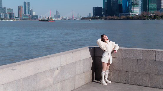 Beautiful young woman with black long hair in white skirt posing eyes closed with blur Shanghai Bund landmark buildings background in sunny day. Emotions, people, beauty, travel and lifestyle concept.
