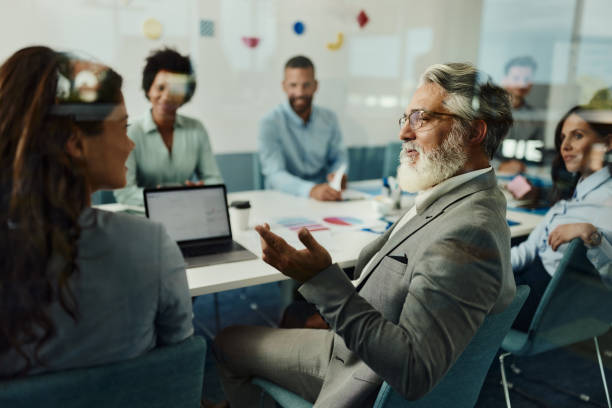 Senior businessman talking to his colleague on a meeting in the office. Mature male entrepreneur communicating with his colleague during a meeting in a modern office. The view is through glass. career vitality stock pictures, royalty-free photos & images