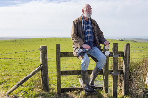 Bearded man in his 70s sitting on a stile in the Winter countryside in Wales, with the sea just visible in the far background.