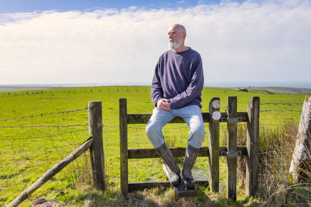 Bearded man in his 70s sitting on a stile in the Winter countryside in Wales, with the sea just visible in the far background.