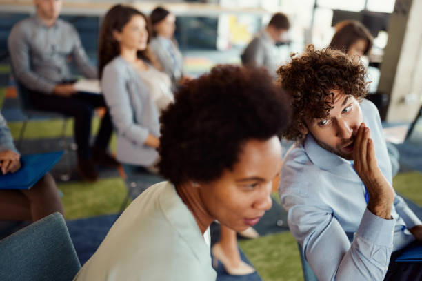 Gossiping on a business seminar! Young male entrepreneur whispering something to his black female colleague during an education event in board room. whisper men stock pictures, royalty-free photos & images