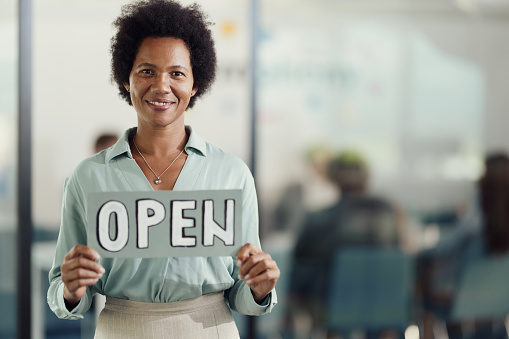 Happy African American businesswoman holding open sign in the office and looking at camera.
