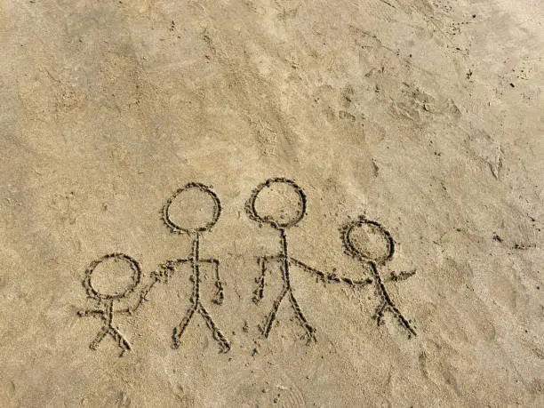 Photo of Close-up image of drawing of stick figure family drawn in damp sand with stick on a sunny beach, parents holding children's hands, siblings, adults not holding hands, elevated view, separation and divorce concept