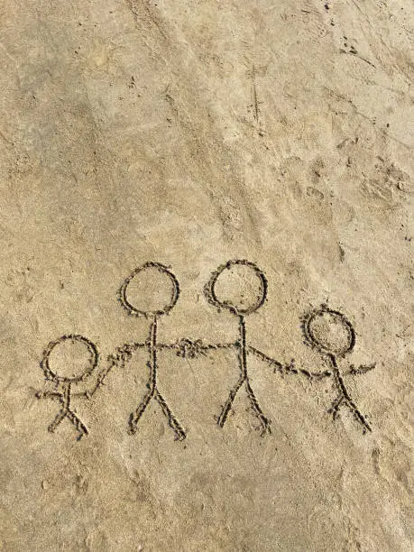 Photo of Close-up image of drawing of stick figure family drawn in damp sand with stick on a sunny beach, parents holding children's hands, siblings, adults holding hands, elevated view, happy families concept, copy space