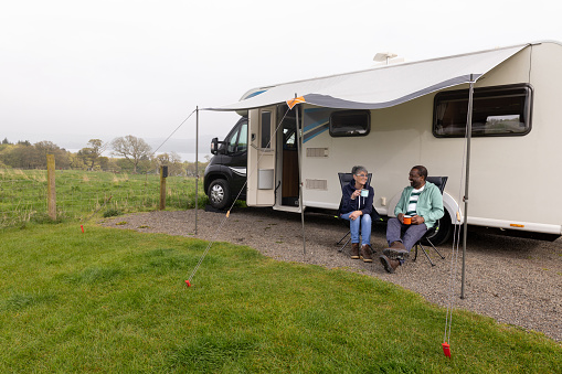 Two senior friends on a hiking staycation, staying in a camper van in Dumfries and Galloway, Scotland. They are sitting outside drinking tea.