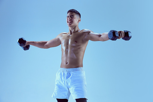 Portrait of young muscular man training with dumbbells, spreading hands isolated over blue background in neon light. Sport, fitness, healthy and active lifestyle, motivation, workout, strength concept