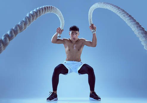 Portrait of young muscular man training, pulling rope isolated over blue background in neon light. Strong hands and legs. Concept of sport, fitness, healthy and active lifestyle, motivation, workout