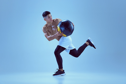 Portrait of young muscular man training, running with med ball isolated over blue background in neon light. Concept of sport, fitness, healthy and active lifestyle, motivation, workout