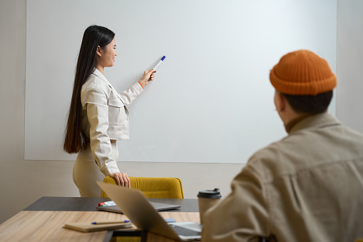 Joyous female office employee pointing with marker at whiteboard to her colleague seated at desk