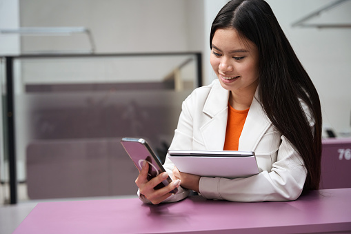 Joyful Asian corporate worker with portable computer in hand looking at cellphone touch screen