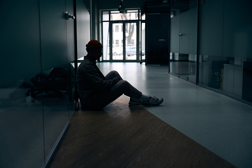 Side view of depressed company employee sitting on floor by glass wall