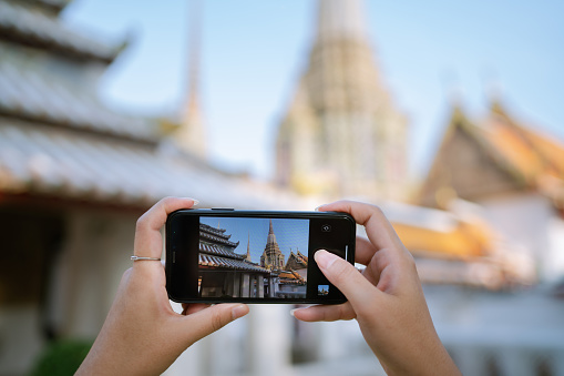 A close-up view of a young female tourist using her mobile phone to take a picture of a beautiful temple during her visit on Bangkok.