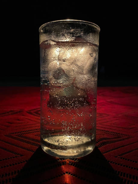 Image of a backlit glass full of ice in a dark room on a red table and a clear fizzy drink, lemonade, gin and tonic, sparkling water, seltzer Stock photo showing a backlit glass full of ice in a dark room on a red table and a clear fizzy drink, it could be alcoholic or a soft drink, such as lemonade, gin and tonic, sparkling water, seltzer fruit garnish stock pictures, royalty-free photos & images