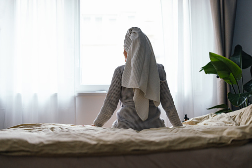 Back view of unrecognizable woman in bathrobe looking through the window while sitting in the bedroom.