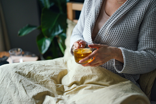 From above photo of an anonymous sick woman in a bathrobe drinking a cup of hot tea while resting in her bed at home.