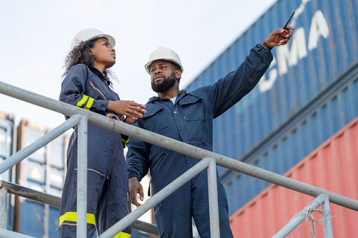 Logistic import and export terminal service make customer satisfaction. African American dock engineers standing on the shipment rack in front of container box and point to shipment cargo in the container warehouse area. Discussing on the stock inventory.