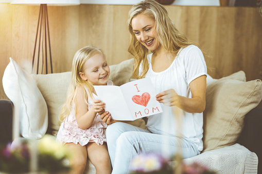 Happy mother day in sunny flat. Child daughter congratulates mom and gives her postcard with heart drawing. Family concept.