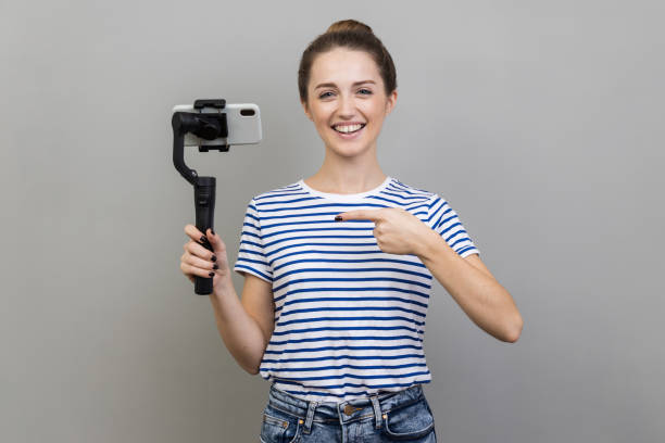 istock Woman holding mobile phone, pointing at steadicam, looking at camera with positive expression. 1474179850