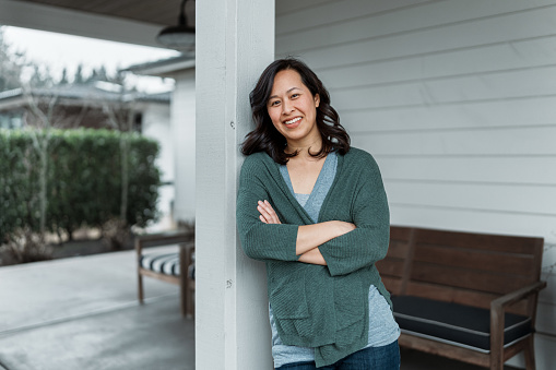 Midrange portrait of a Vietnamese woman smiling confidently at the camera while leaning casually against a pillar on the covered front patio of her modern farmhouse style home.