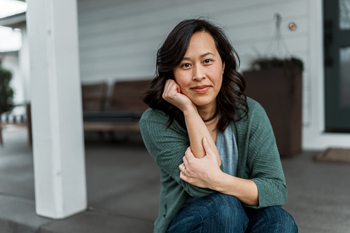 Portrait of a beautiful Vietnamese woman with a contemplative expression looking directly at the camera while sitting on the step leading to the covered patio and front door of her home.