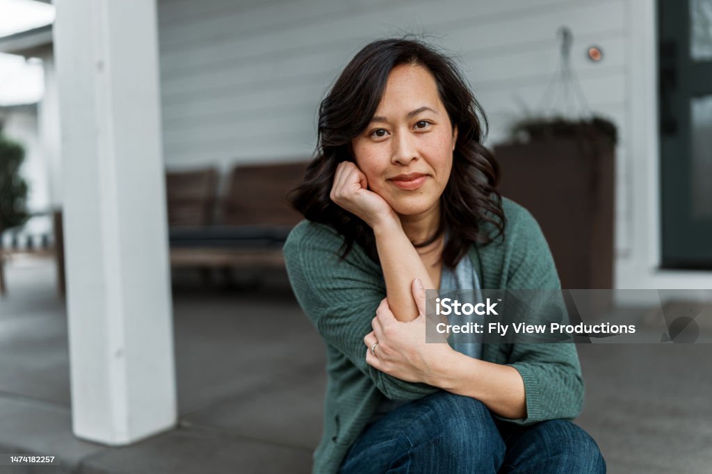 Woman sitting on front step of home Portrait of a beautiful Vietnamese woman with a contemplative expression looking directly at the camera while sitting on the step leading to the covered patio and front door of her home. Portrait Stock Photo