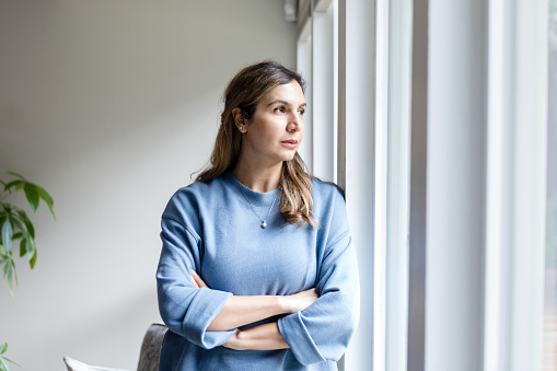 The serious mid adult businesswoman stands with her arms crossed as she looks out her office window.