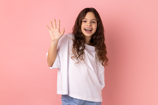 Portrait of friendly positive little girl wearing white T-shirt smiles toothily, raises palm greets friend, saying hello or goodbye. Indoor studio shot isolated on pink background.