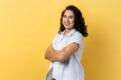 Portrait of satisfied delighted happy woman with dark wavy hair standing with folded hands and looking at camera with toothy smile. Indoor studio shot isolated on yellow background.