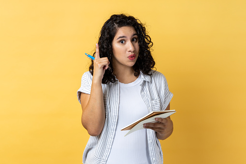 Portrait of amazed inspired woman journalist with dark wavy hair writing in paper notebook, having new excellent idea, raising finger up. Indoor studio shot isolated on yellow background.