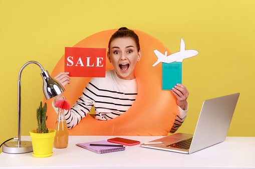 Excited surprised woman with rubber ring, holding card with sale inscription, passport and paper plane, amazed with summer sales for tours. Indoor studio studio shot isolated on yellow background.