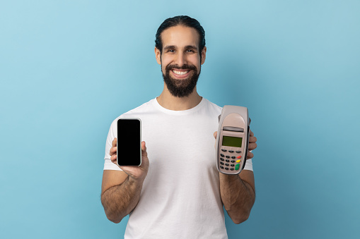 Portrait of smiling happy man with beard wearing white T-shirt holding pos terminal and smart phone with empty display for advertisement. Indoor studio shot isolated on blue background.