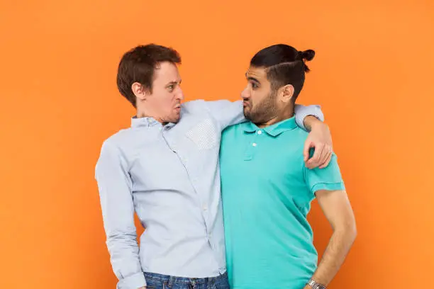 Portrait of two comic men friends standing and hugging, looking at each other with funny frowning facial expressions. Indoor studio shot isolated on orange background.
