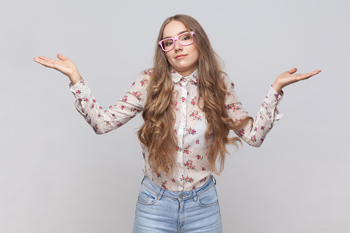 I don't know. Portrait of confused beautiful woman in glasses with wavy blond hair standing, raised arms, confused and thinking. Indoor studio shot isolated on gray background.
