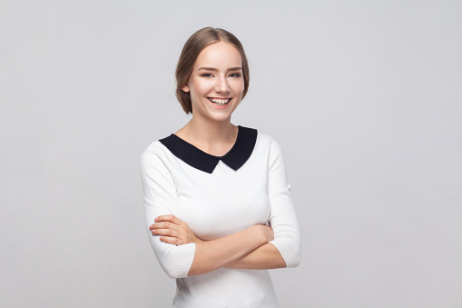 Portrait of smiling friendly woman wearing white dress standing with crossed arms, looking at camera with toothy smile, expressing positive emotions. Indoor studio shot isolated on gray background.