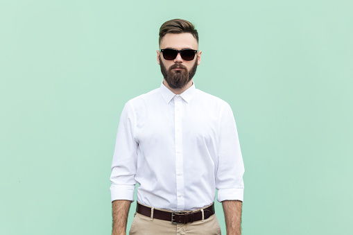 Portrait of handsome bearded businessman wearing white shirt and black sunglasses standing with serious and confident facial expression. Indoor studio shot isolated on light green background.