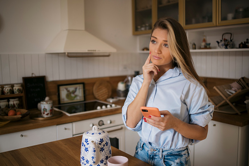 Thoughtful young woman holding smart phone while standing at the kitchen counter. Caucasian female thinking while reading text message on her mobile phone at home.