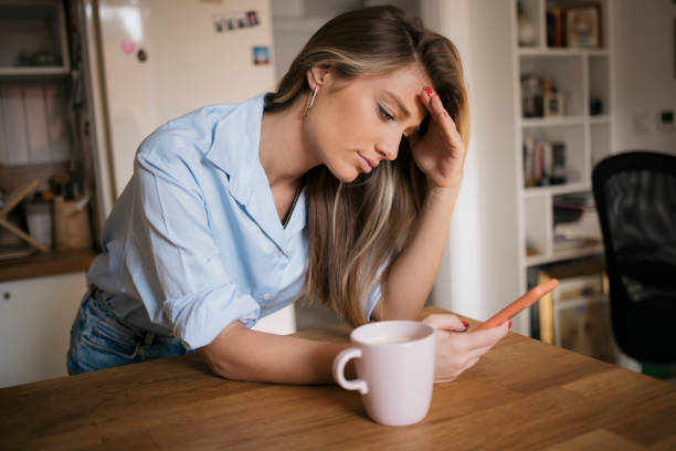 Young woman using her cellphone while drinking coffee in kitchen stock photo