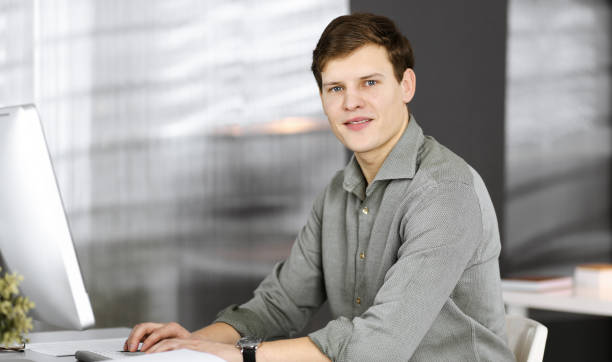 istock Young businessman and programmer in a green shirt is looking at camera, while he is working on his computer, sitting at the desk in a cabinet. Headshot or business portrait in an office 1474188225