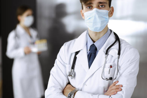 jeune docteur mâle restant avec des bras croisés dans la clinique. médecin intelligent utilisant le masque médical protecteur dans l’hôpital moderne. concept de médecine et de virus corona - clothing professional occupation medical occupation doctor photos et images de collection