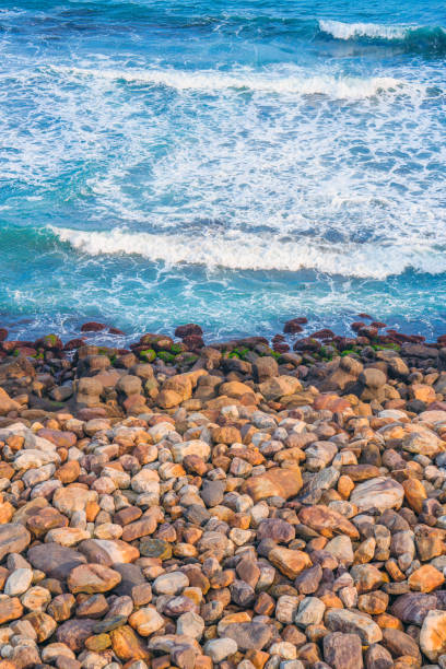 The coastal view of yellow-orange rocks lining the shore with the blue sea and white rolling waves crashing onto the beach. The contrasting colors of the rocks and sea make for a striking and beautiful scene.