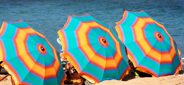 Three beach umbrellas in front of the sea, sunbathing underneath. Beach sand and sea water in the background.  Rías baixas, Pontevedra province, Galicia, Spain.