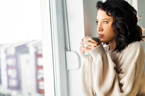 istock Young woman relaxing at home drinking coffee looking though window 1474192927