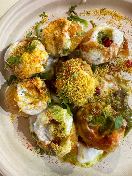 Stock photo showing elevated view of white plate containing golgappa puri snacks being served filled with boiled potato, onion, pomegranate seeds, and spicy chutneys, topped with herbs, spices, yoghurt and Sev (crunchy noodles). imagen de golgappa puri (bolas de agua fritas), esferas huecas crujientes con agujeros, relleno de papas, cebolla, semillas de granada y chutneys picantes, dahi papri chaat, cubierto con hierbas y especias, yogur, sev (fideos crujientes), vista elevada