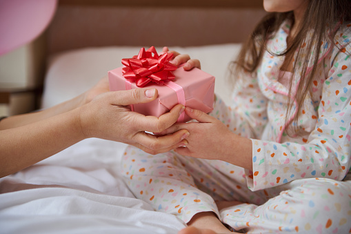 Selective focus on hands of woman and child, holding gift box wrapped in pink wrapping paper and tied with red bow. Mother gives a happy present to her lovely daughter for her birthday party. Close-up