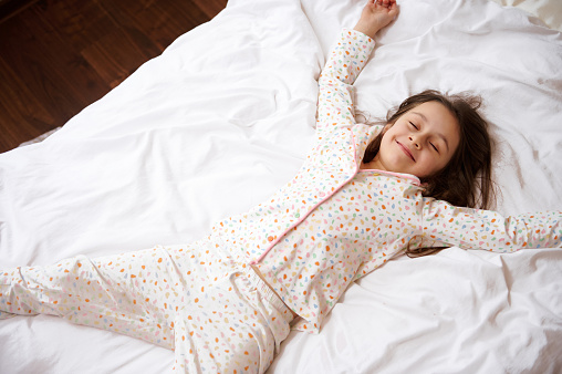 View from above of an adorable little child girl with closed eyes, in stylish cotton pajamas with colorful dots, smiling broadly while lying down on a comfortable bed with orthopedic mattress